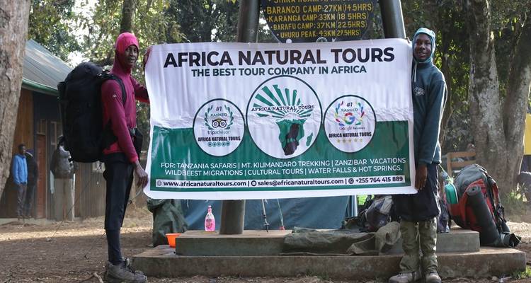 Two hikers pose with Kilimanjaro trekking company banner near forest camp
