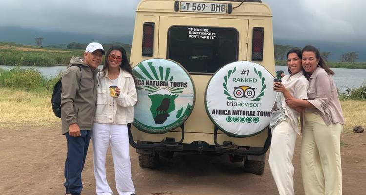 Group poses beside a safari jeep with tour company spare-wheel covers near a lake and cloudy hills