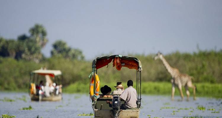 Botes turísticos en un río tranquilo con una jirafa lejana parada en agua poco profunda