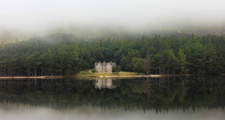 Mistig zicht op een statig landhuis omringd door bomen weerspiegeld in een meer.