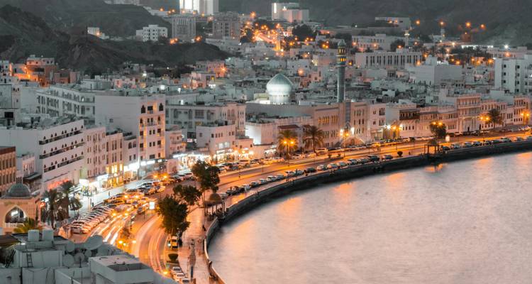 Vista elevada de la carretera costera curva de Muscat iluminada por farolas doradas al atardecer.