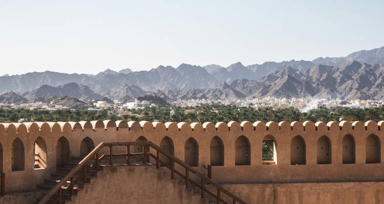 Muro almenado de un fuerte desértico que domina un oasis de palmeras y una cadena montañosa escarpada.