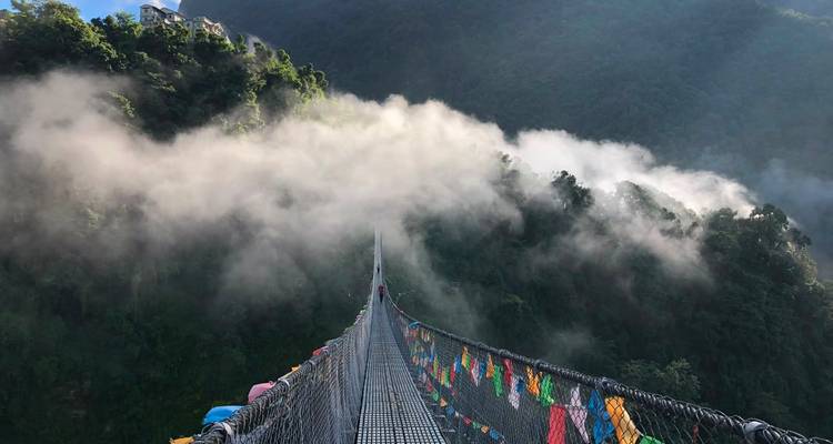 A long suspension bridge draped with prayer flags disappears into mist over a lush Nepali gorge.