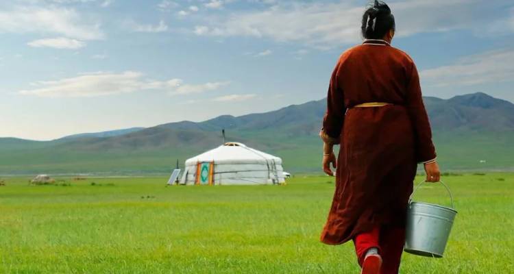 Eine Mongolin in traditioneller Deel trägt einen Metalleimer über die weite grüne Steppe zu einer entfernten Jurte mit Bergen im Hintergrund.
