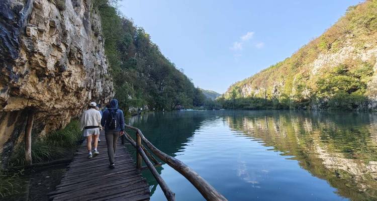 Twee mensen die wandelen op een houten pad langs een serene meer omringd door kliffen en groen.