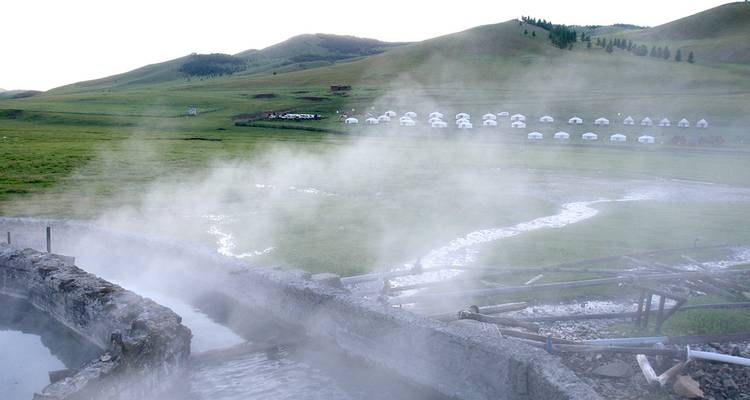 Steam rises from natural hot-spring pools on a grassy Mongolian plain with ger camp in distance.