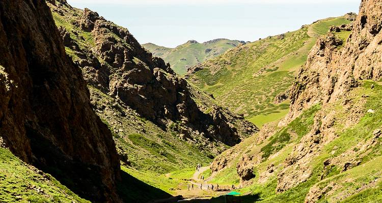 A narrow green valley framed by steep rocky cliffs with small hikers visible on the trail below.