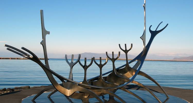 Künstlerische Metallskulptur am Meer mit Bergen im Hintergrund.