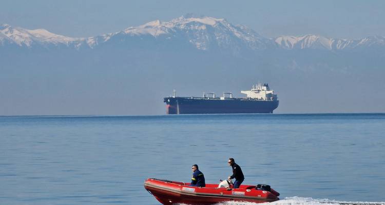 Two people speed across calm blue sea in a red inflatable boat with a large cargo ship and snowy mountains in the distance.