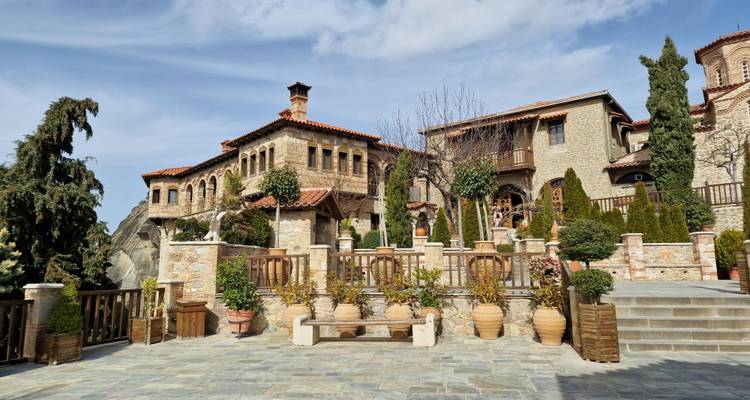 Stone monastery buildings with terracotta roofs, potted plants and carved balconies at Meteora under blue sky.