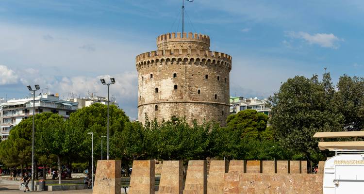 The historic White Tower of Thessaloniki rises behind leafy trees and modern city elements on a sunny day.