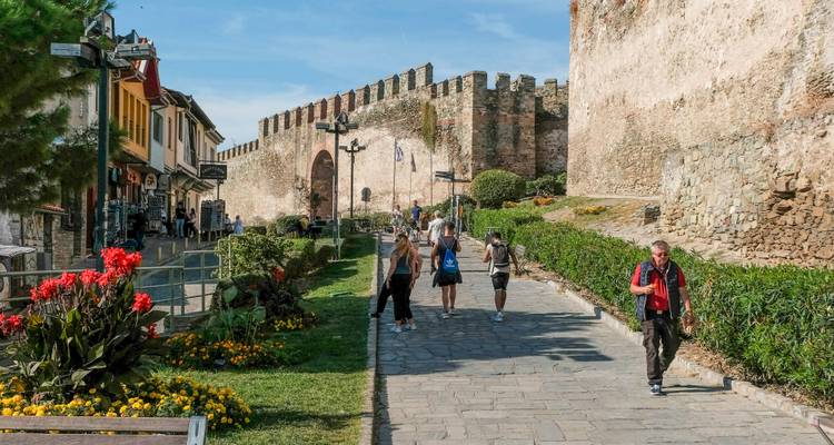 Visitors walk along a cobbled path beside historic stone walls and vibrant flowerbeds in Thessaloniki’s Upper Town.