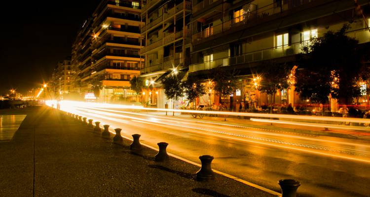 Long-exposure streaks of car lights along Thessaloniki’s nighttime waterfront lined with cafes and apartments.