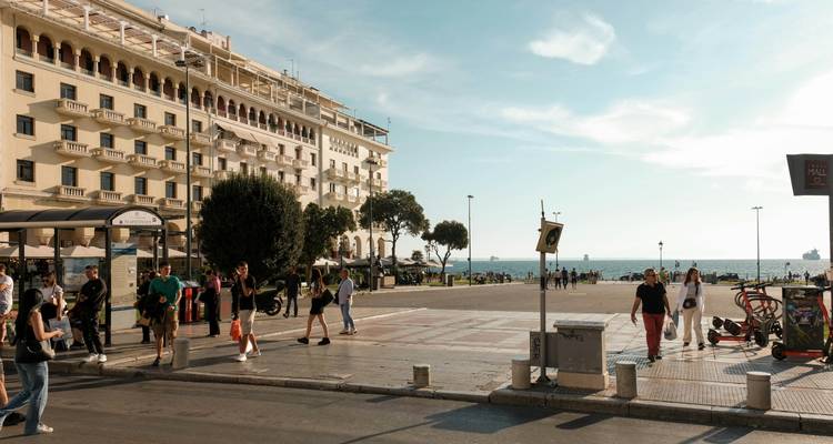 Seaside promenade in Thessaloniki with people walking, cafes and views toward the open sea.
