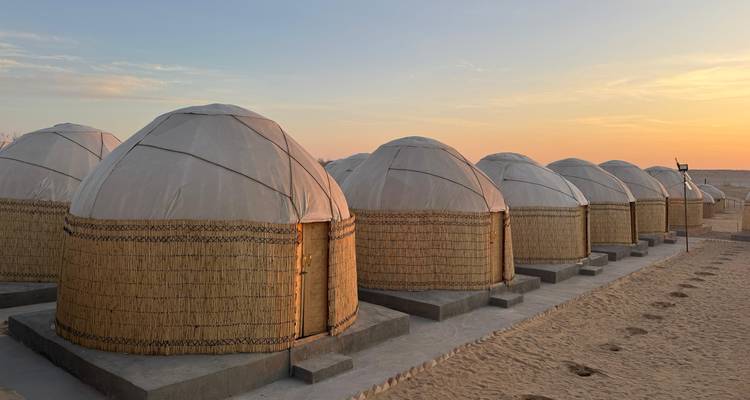 Row of traditional yurts lined up on sandy desert ground under a warm sunset sky.