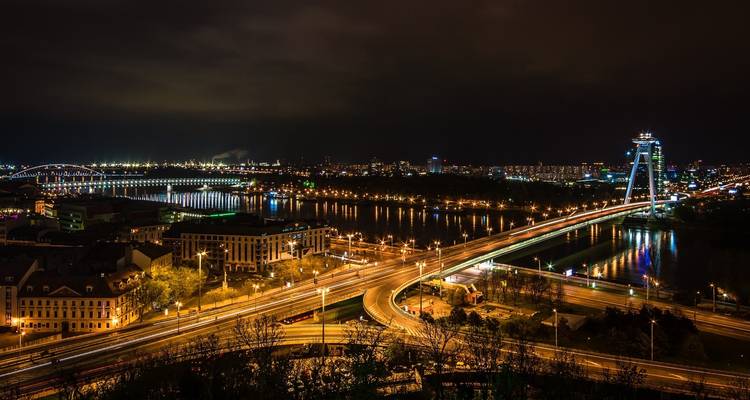Panorama nocturno de Bratislava con puentes iluminados y luces de la ciudad reflejándose en el Danubio.