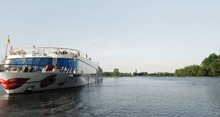 Ein modernes weißes Flusskreuzfahrtschiff mit rotem Lippendesign gleitet über einen ruhigen, von Bäumen gesäumten Fluss unter klarem Himmel.