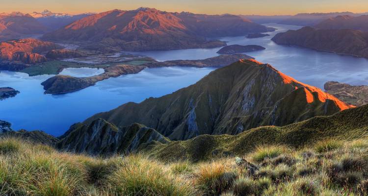 Golden morning light washes over Roys Peak ridgeline above Lake Wanaka and a maze of blue inlets and distant alpine ranges.