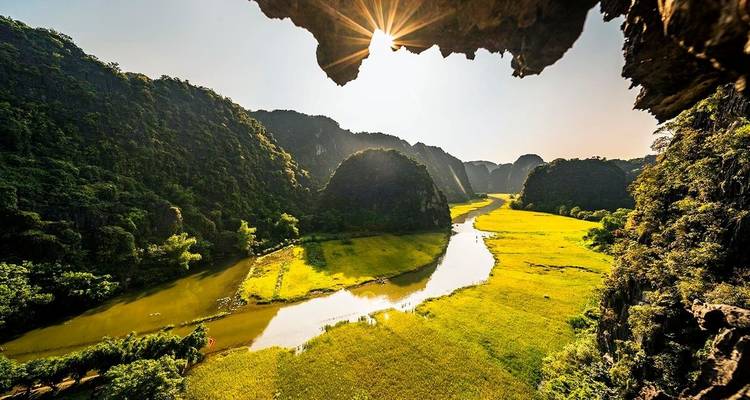 Scenic view of a river winding through lush green fields with mountains in the background and sunlight peeking through rocks.