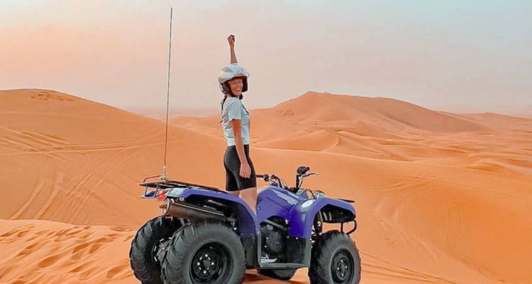 Avonturier op een paarse quad die triomfantelijk zijn vuist omhoogsteekt bovenop uitgestrekte oranje zandduinen.