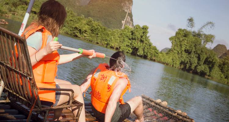 Deux voyageurs en gilets de sauvetage s'éclaboussent mutuellement avec des pagaies sur un radeau en bambou dans une rivière pittoresque.