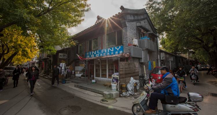 Ruelle hutong étroite avec des habitants marchant et circulant à scooter, la lumière du soleil inondant les bâtiments en briques grises.