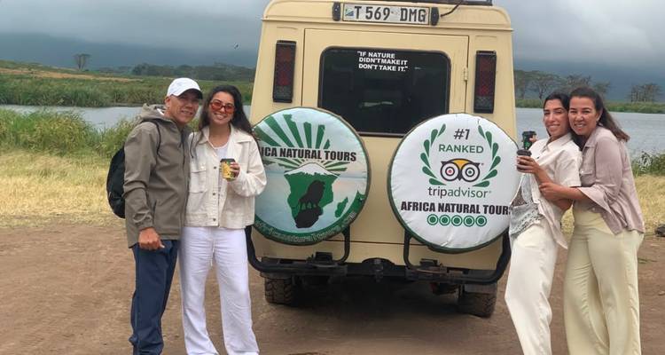 Travellers pose beside safari vehicle backed by a lake and cloudy sky.