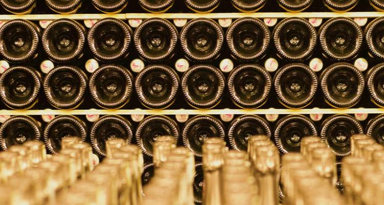 Filas de botellas de vino envejecidas apiladas horizontalmente en una bodega tenue con iluminación dorada cálida.