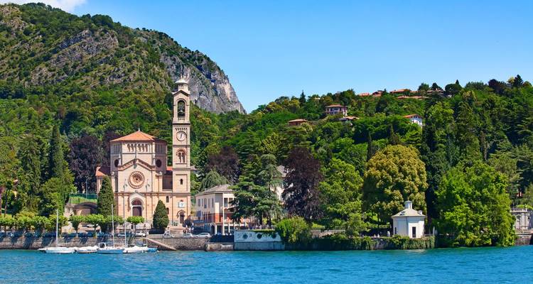 Iglesia histórica junto al lago con una torre de campanario alta rodeada de vegetación exuberante y aguas azules bajo un cielo despejado.