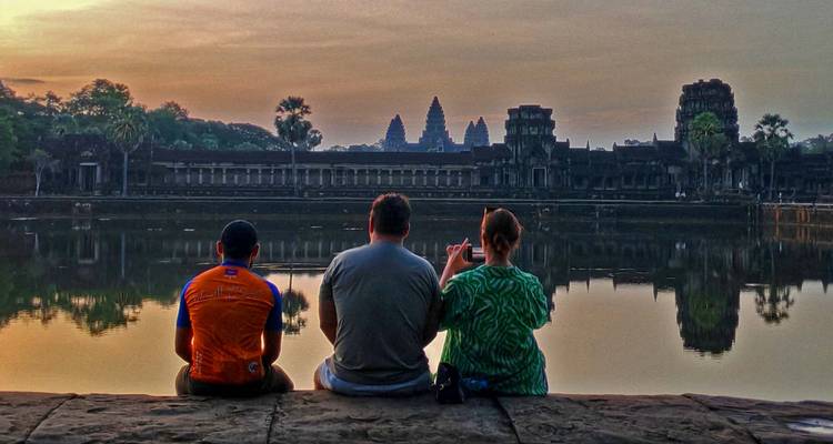 Los viajeros se sientan junto al lago espejado observando el amanecer sobre la silueta de Angkor Wat