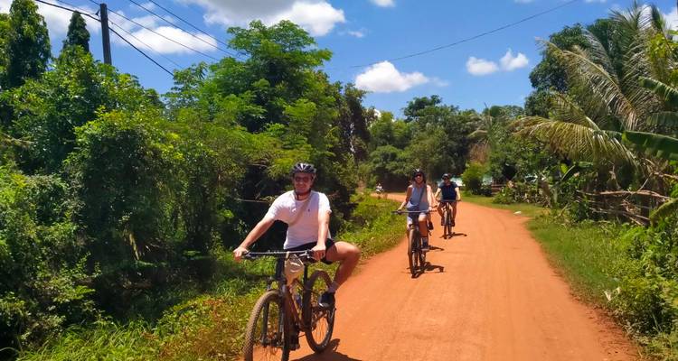Los ciclistas bajan por un camino de tierra roja rodeado de vegetación densa bajo un cielo soleado.