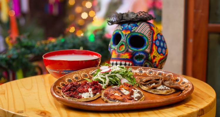 Mexikanische Tacos und bunte Totenkopfdekoration auf einem Holztisch rufen die Atmosphäre des Día de Muertos hervor.