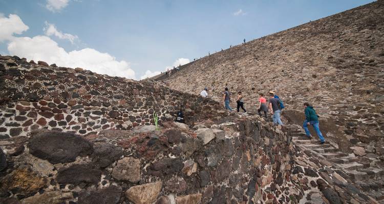 Reisende erklimmen steile Steinstufen der Teotihuacán-Pyramide vor blauem Himmel.