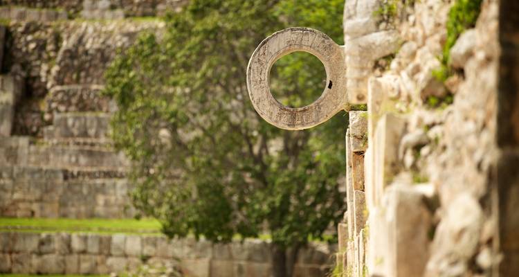 Antiker Steinring eines Maya-Ballplatzes steht gegen verschwommene Ruinen und Grünflächenauf.