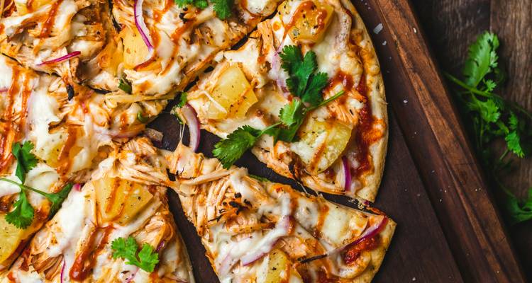 Wooden board pizza topped with shredded chicken, pineapple, red onion, cheese and cilantro drizzled with sauce.
