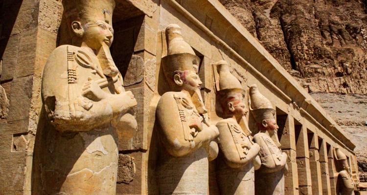 Een rij standbeelden die oude Egyptische figuren uitbeelden, staand tegen een rotsachtige achtergrond.