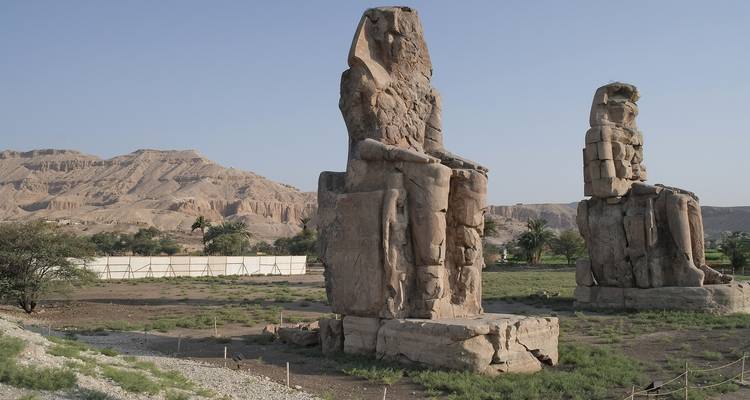 Twee grote oude stenen beelden in een woestijnlandschap met bergen op de achtergrond.