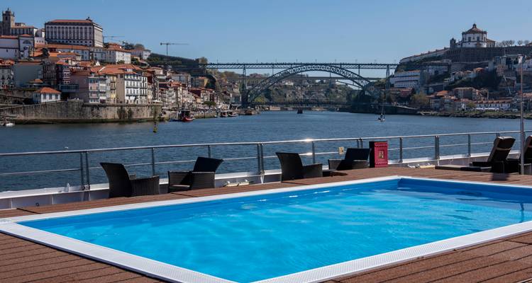 Ein Pool auf einem Schiffsdeck mit Blick auf einen Fluss, eine Brücke und eine Stadtlandschaft.