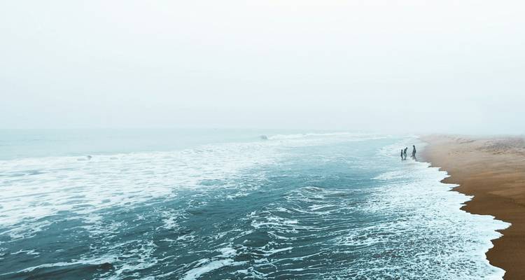 Nebliger Strand mit sanften Wellen und entfernten Gestalten, die spazieren gehen.