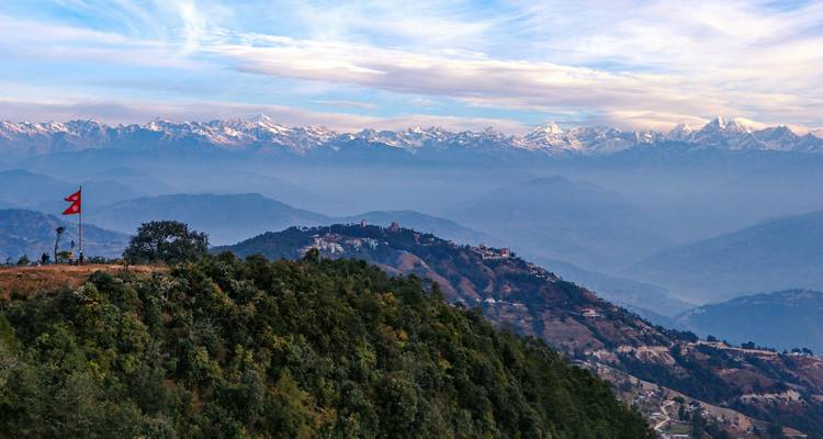 Vista panorámica de colinas frondosas y montañas nevadas distantes con una bandera, posiblemente en Nagarkot, Nepal.