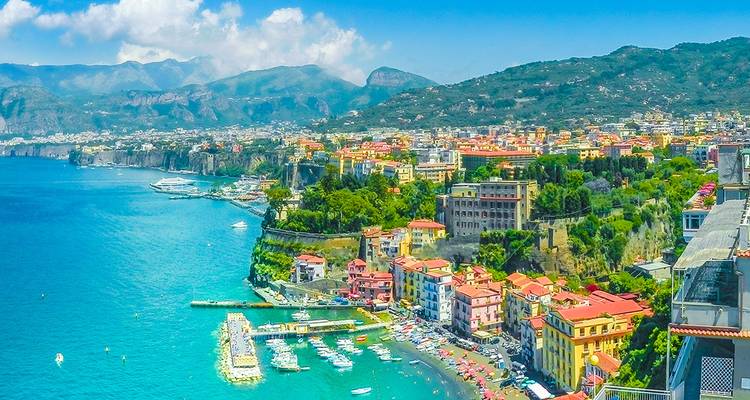 Un panorama costero amplio muestra el pueblo de Sorrento en lo alto del acantilado, el mar turquesa, el puerto deportivo y las montañas distantes bajo un cielo brillante.