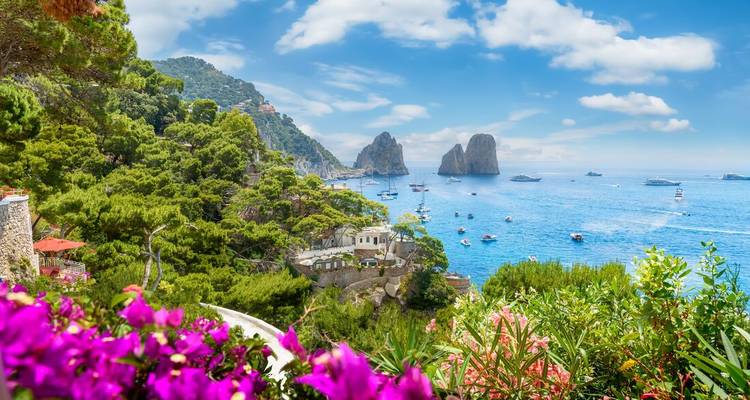 Flores brillantes enmarcan una vívida vista costera de las formaciones rocosas Faraglioni de Capri y barcos flotando en agua turquesa.