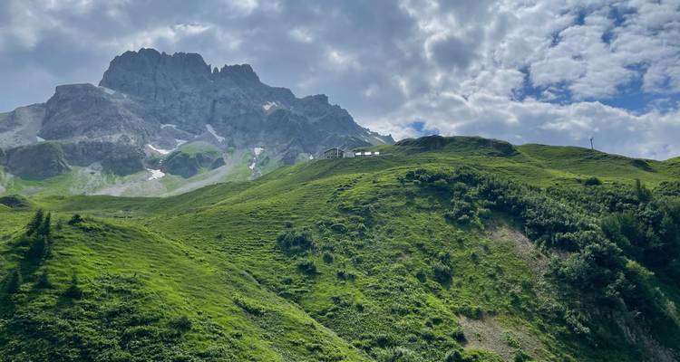 Des pentes alpines verdoyantes mènent à un sommet gris dentelé sous un ciel partiellement nuageux.