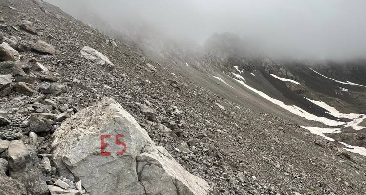 Pente de éboulis rocailleux et brumeux avec marque de sentier E5 rouge sur la pierre et plaques de neige.