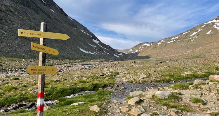 Des panneaux de signalisation alpins jaunes se dressent dans une vallée de montagne rocheuse avec des plaques de neige fondue et un cours d'eau.