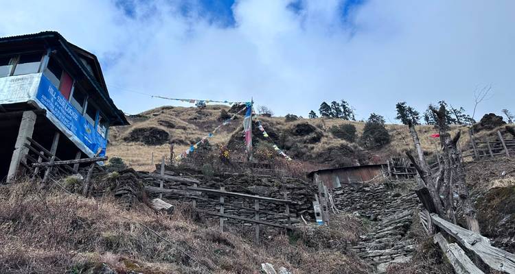 Mountain village buildings and prayer flags on a dry hillside under partly cloudy sky in Nepal.