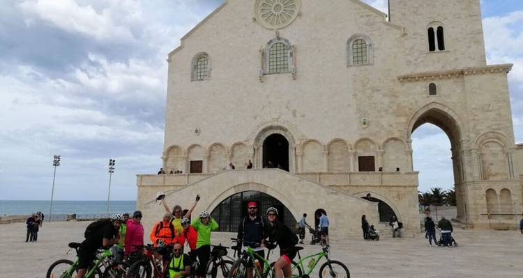Cyclists with bicycles gathered in front of a large white historical building near the sea.