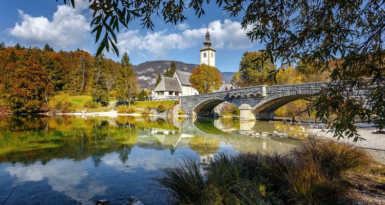 A stone bridge and white church reflect in the crystal clear waters of Lake Bohinj amid autumn foliage.