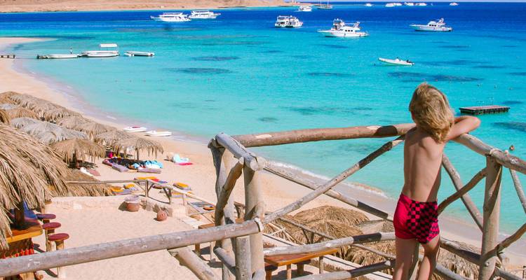 Repeat scene with child overlooking pristine turquoise bay and sandy beach from wooden deck.