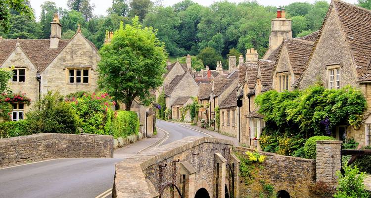 Casitas de piedra con encanto y verdor exuberante bordean una carretera sinuosa a través de un pueblo clásico de Cotswolds con un pequeño puente de piedra en primer plano.